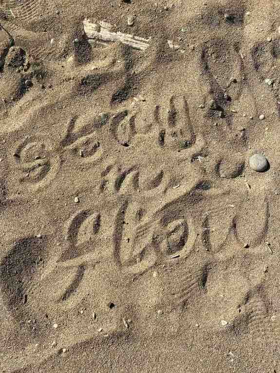 Words written in cursive on sandy beach ground read 'stay in flow,' surrounded by footprints and small pebbles — a grounding reminder of unwind rituals.