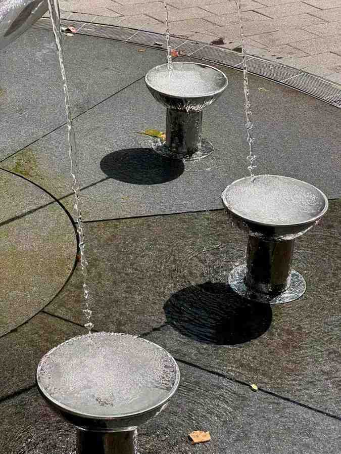 Streams of water gently arch into three shallow metal basins of a fountain, creating ripples and reflections on the dark stone ground — a calming reminder of soft flow.