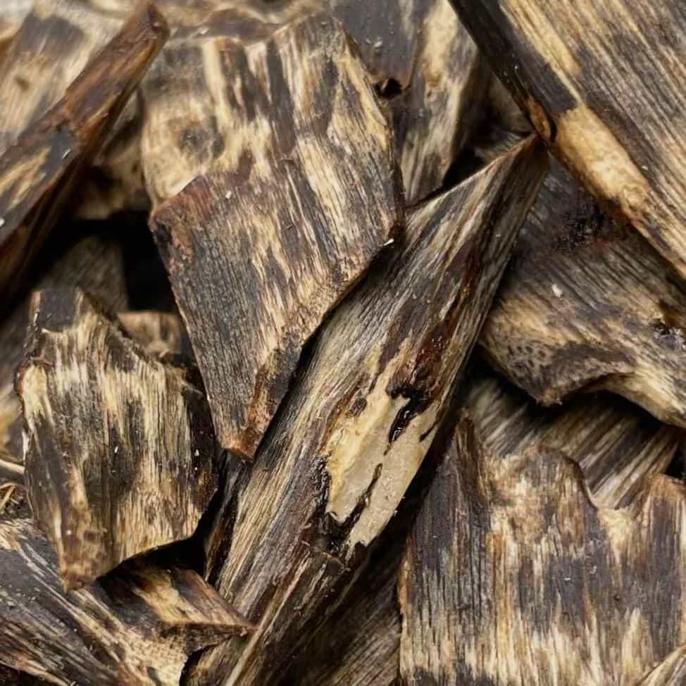 A close-up of a pile of agarwood chips, revealing their distinct dark and light grain patterns and rough, splintered edges. The image represents a scent that is deep and resinous, and has been treasured in meditation for its ability to still the mind, sharpen awareness, and create a sacred atmosphere.