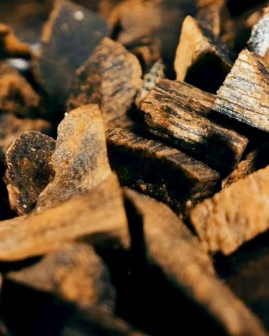 A close-up of a pile of agarwood chips, revealing their distinct dark and light grain patterns and rough, splintered edges. The image represents a scent that is deep and resinous, and has been treasured in meditation for its ability to still the mind, sharpen awareness, and create a sacred atmosphere.
