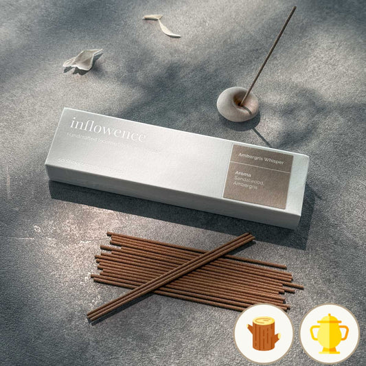 Inflowence Ambergris Whisper incense box with sandalwood and ambergris incense sticks neatly arranged beside a lit stick in a ceramic holder—an aromatic ritual designed to support emotional reset and grounding.