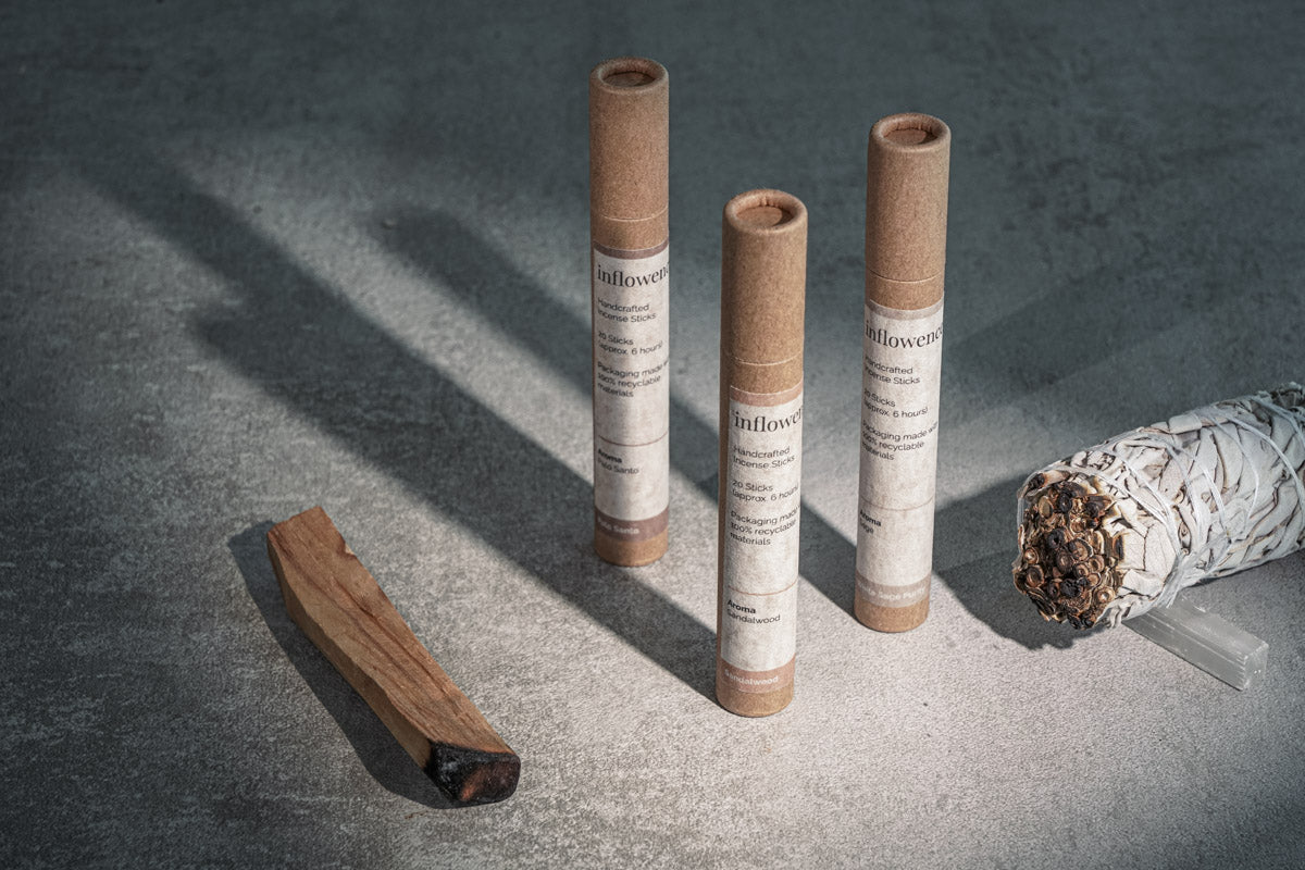 Three cylindrical kraft paper tubes labeled 'inflowence' handcrafted incense sticks standing upright on a stone surface, alongside a white sage smudge bundle and a partially burned palo santo stick, with soft shadows cast across the scene.