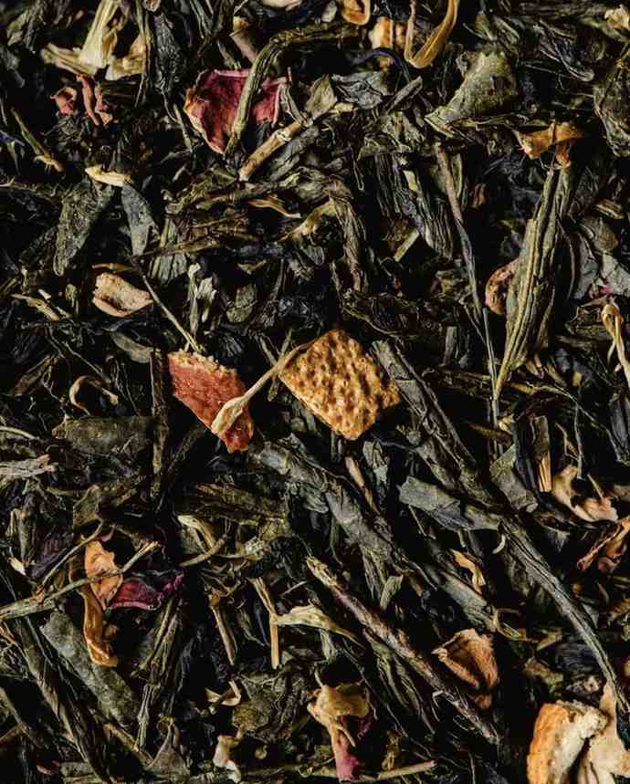 A close-up, top-down view of a textured mix of dried green tea leaves, dried fruit pieces (possibly citrus), and dark red petals, creating an aromatic blend. The image represents herbs that positively influence Mood & Emotion, enhance Mind & Focus, and contribute to a desired Atmosphere & Practice.