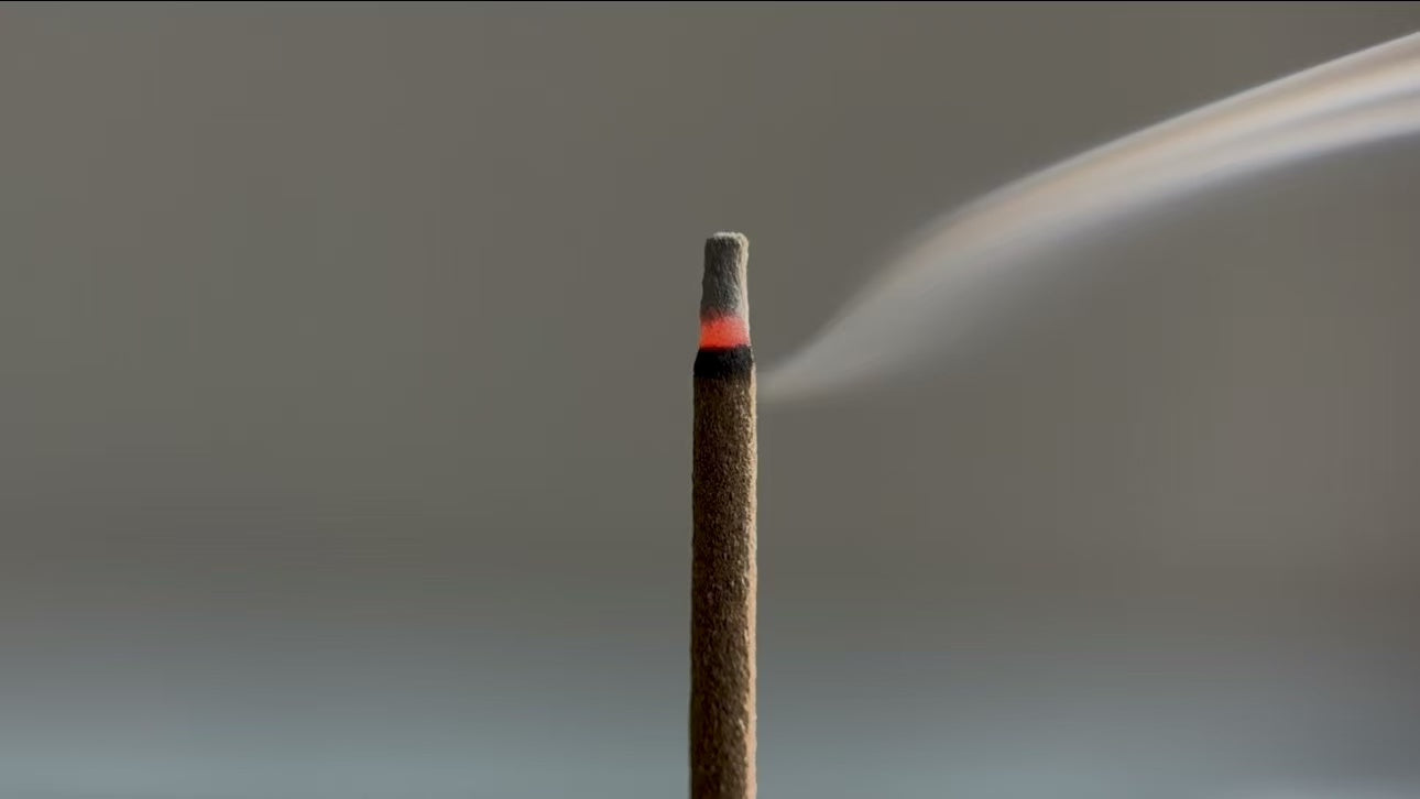 A close-up shot of an inflowence incense stick burning, with a glowing red tip and a light wisp of smoke rising from it. The image represents a natural product with a clean and consistent burn.