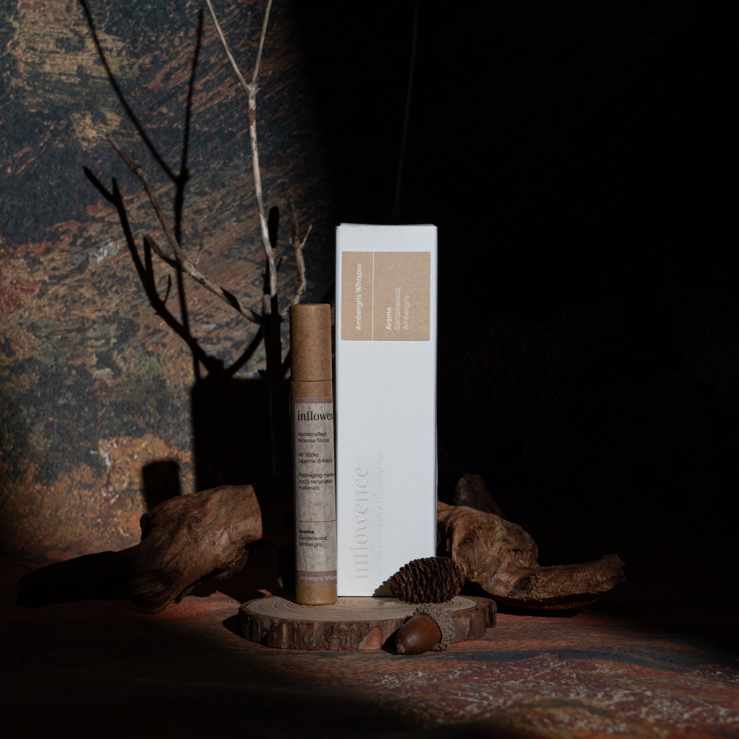Ambergris Whisper incense box and tube set displayed with wood and branches, in front of a green-yellow-red oil painting canvas. Dappled light and shadow highlight the products, evoking a warm, tranquil evening ritual.