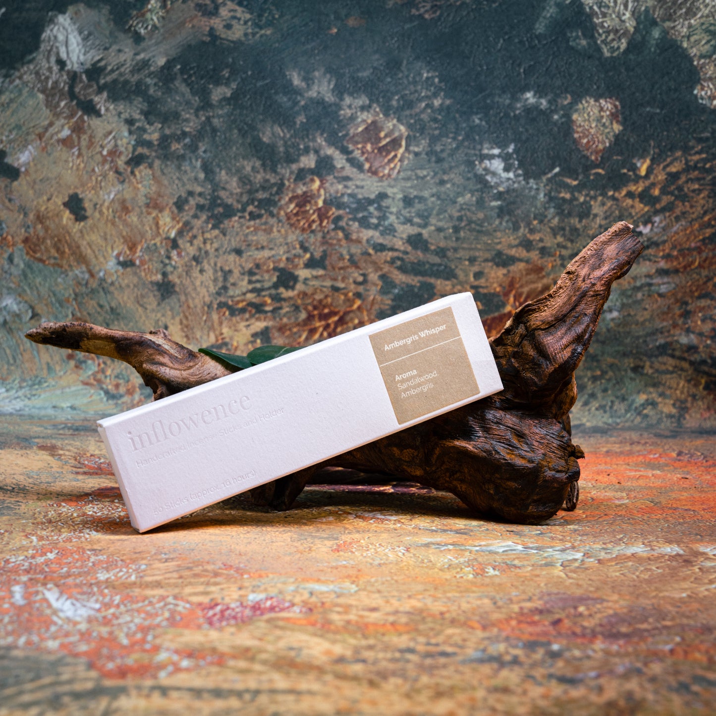 Ambergris Whisper incense box styled with natural wood and greenery, set against a green, red, and yellow oil painting–inspired backdrop — evoking a warm, earthy ritual atmosphere.
