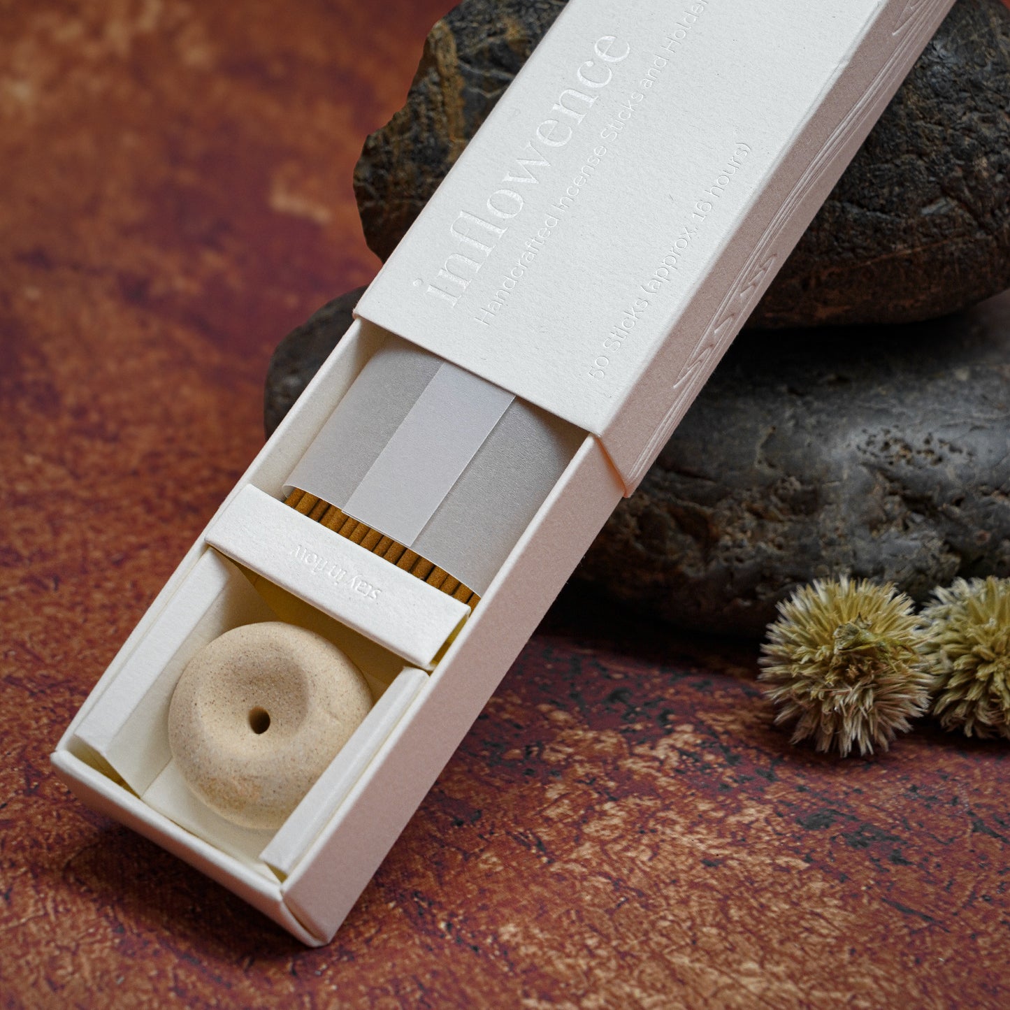 Close-up shot of an open incense box showcasing the packaging, incense sticks, and a clay incense holder, all arranged on a stone surface with a natural textured background.