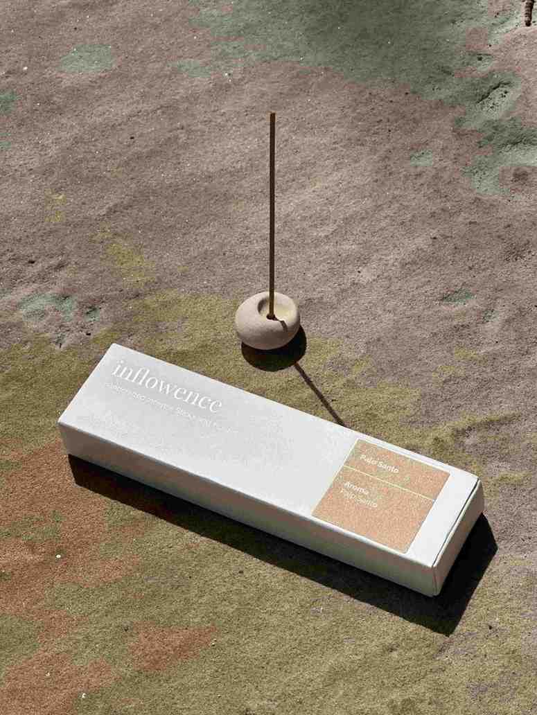 A white inflowence Palo Santo incense box, with embossed text, floating against a light-colored background. The product is designed to reduce pain and improve stress response.
