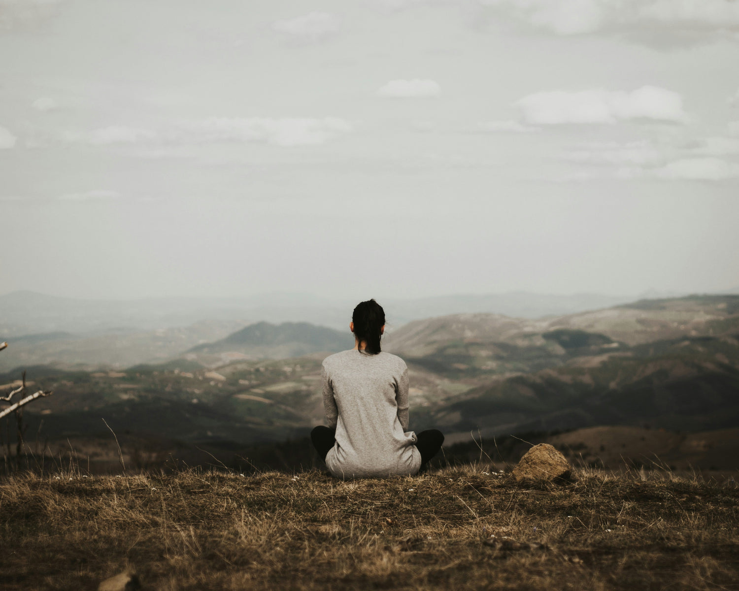 Person sitting cross-legged on the soil, facing a scenic view of rocks and mountains under a cloudy sky—capturing a quiet moment of meditation and connection with nature.