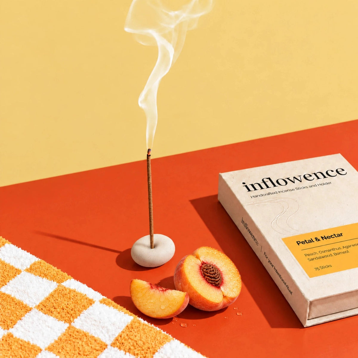 A burning incense stick in a small round holder beside fresh peach halves and a box of Inflowence Petal & Nectar charcoal-free incense of 75 sticks, on an orange and yellow background with an orange checkered rug.