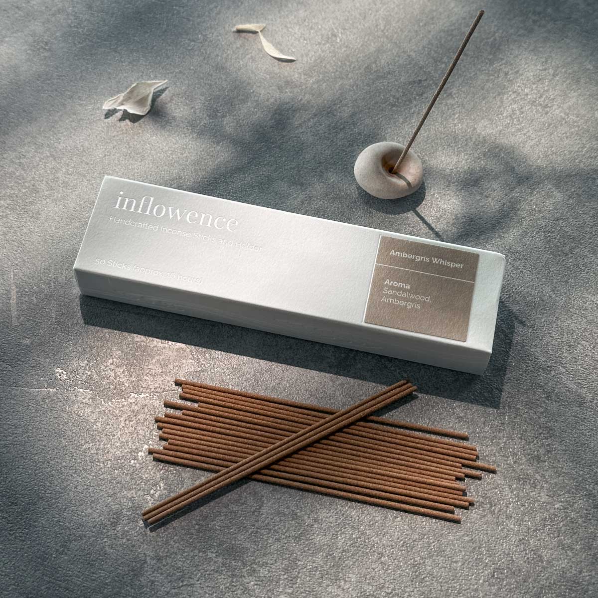 Inflowence Ambergris Whisper incense box with sandalwood and ambergris incense sticks neatly arranged beside a lit stick in a ceramic holder—an aromatic ritual designed to support emotional reset and grounding.