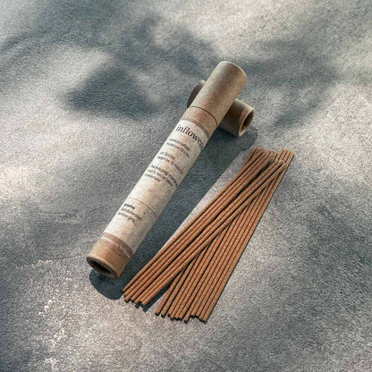 A kraft paper tube of inflowence Ambergris Whisper incense sticks laid beside a neat row of incense sticks on a sun-dappled stone surface—infused with sandalwood and ambergris to create space for emotional reset through mindful rituals.