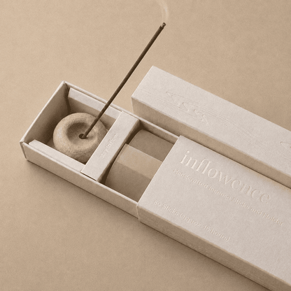 Close-up of an open inflowence incense box with a ceramic holder and a lit incense stick on a brown background.