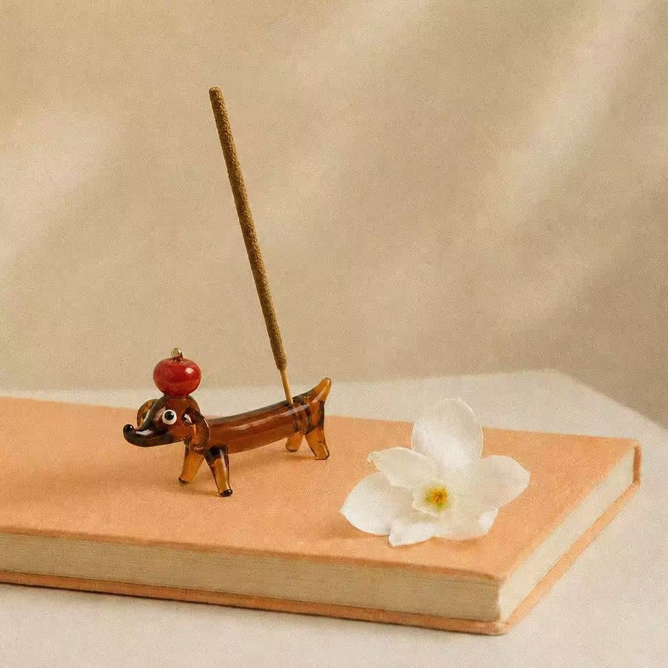 Apple Pup Incense Holder shown on a peach-colored book with a white flower, featuring a hand-shaped glass incense holder in the playful silhouette of a small dachshund balancing a red apple on its head and holding an incense stick.