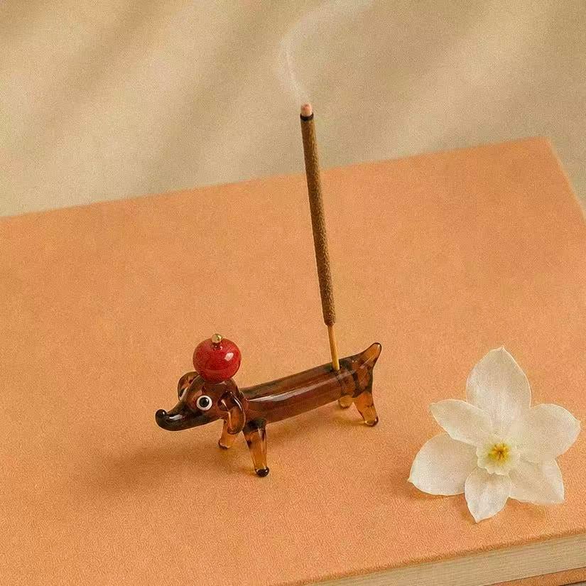 Apple Pup Incense Holder styled on a peach book with a white flower and soft smoke, featuring a playful glass dachshund incense holder balancing a red apple on its head while holding a burning incense stick.