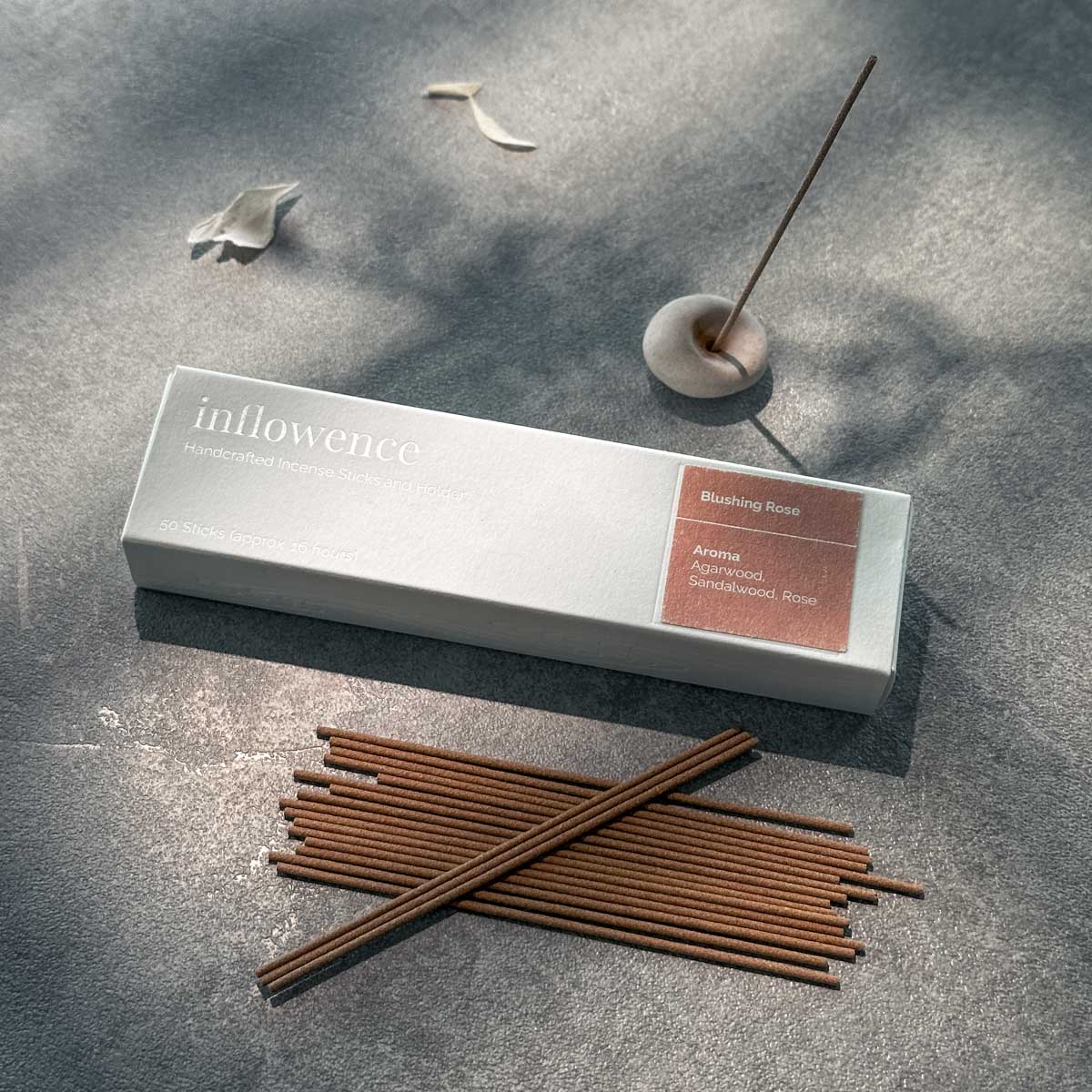 Inflowence Blushing Rose incense box, featuring a soft white design with rose-toned label, accompanied by a lit incense stick in a clay holder and rows of incense sticks on a stone surface—crafted to inspire self love and emotional grounding.
