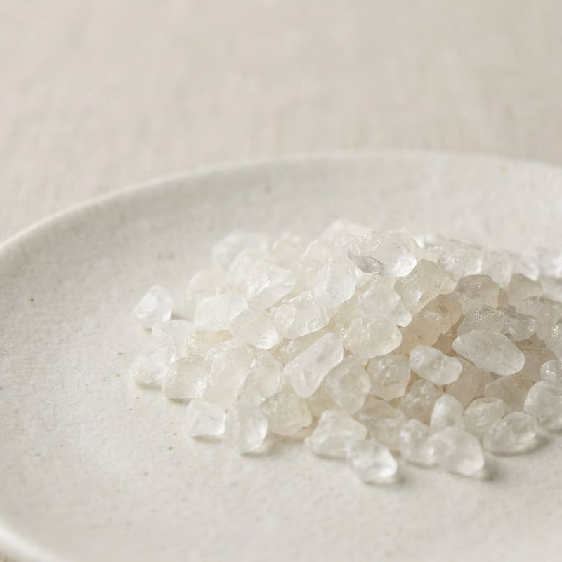 translucent white borneol crystals scattered on a smooth ceramic plate in soft neutral lighting, a key ingredient used in inflowence flower incense named 'Petal & Nectar'.