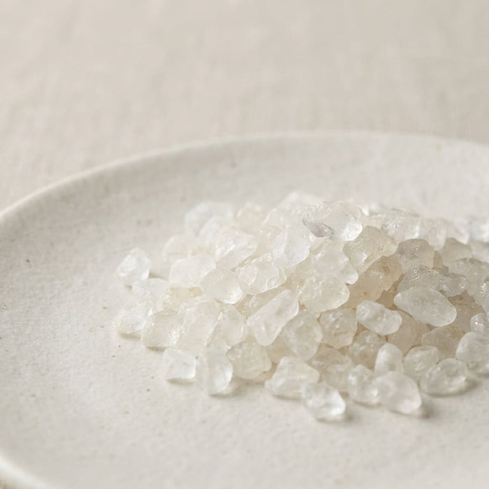 translucent white borneol crystals scattered on a smooth ceramic plate in soft neutral lighting, a key ingredient used in inflowence flower incense named 'Petal & Nectar'.