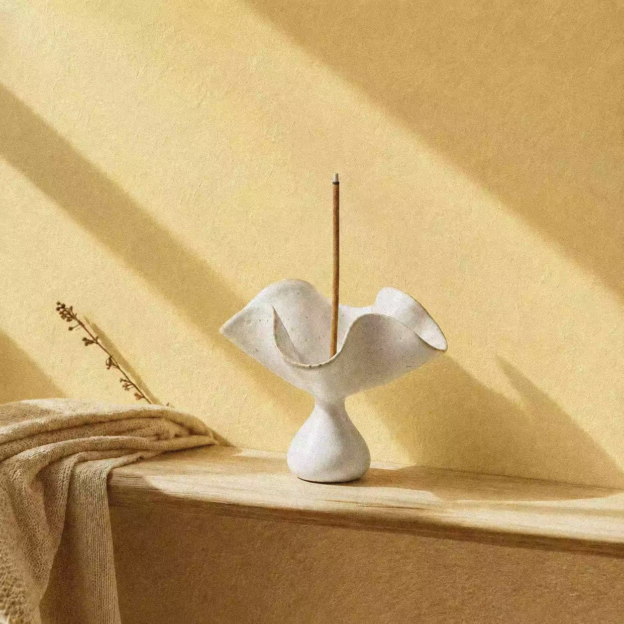 Ceramic incense holder in a sculptural petal shape, shown with a vertical incense stick on a warm wooden surface, soft shadows, and a peaceful daily ritual atmosphere.