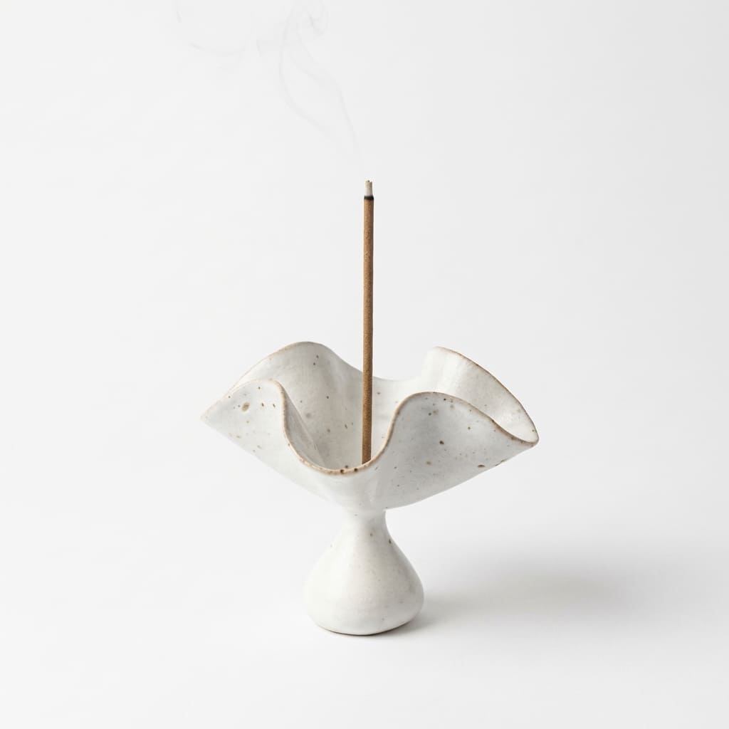 Ceramic incense holder in a hand-formed petal shape, shown on a clean white background with a burning incense stick and soft smoke, designed to hold incense sticks and catch falling ash.