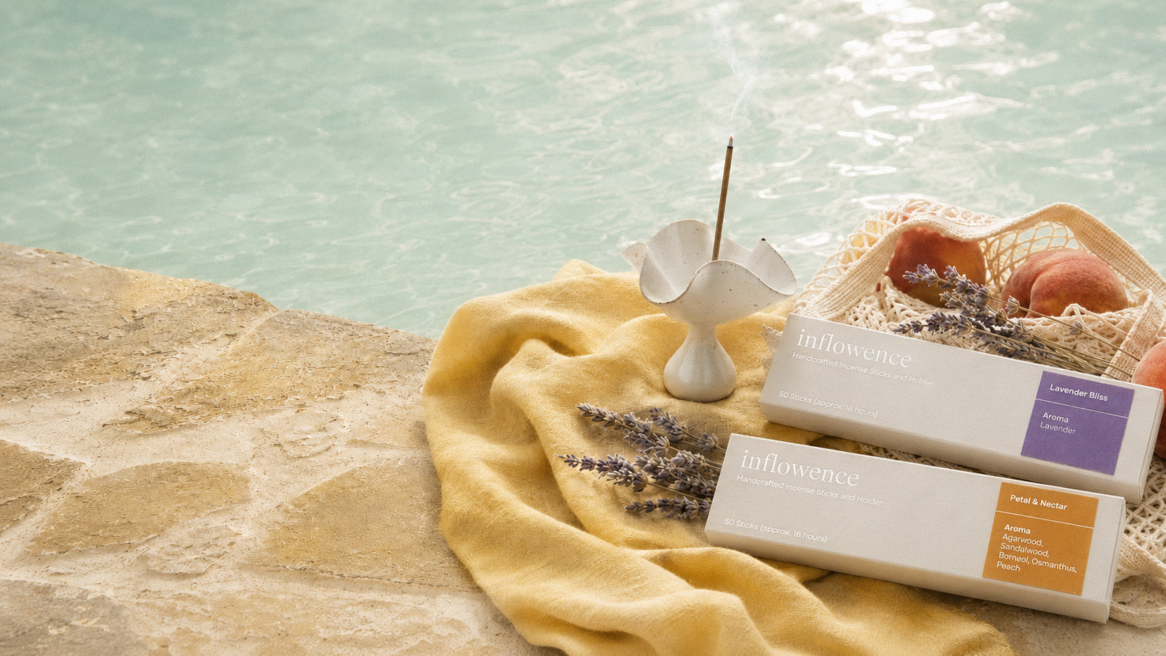Inflowence Lavender Bliss lavender incense box and Petal & Nectar flower incense box displayed beside a pool on soft yellow fabric, with a white petal ceramic incense holder holding a burning incense stick, lavender sprigs, peaches in a woven net bag, textured stone, water reflections, and warm summer light.