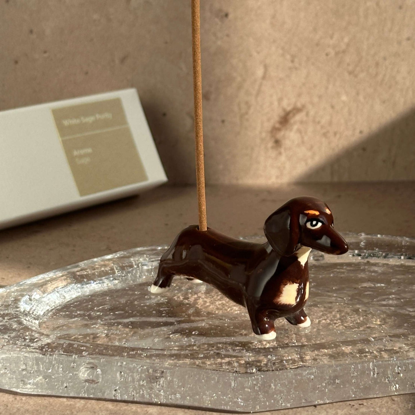 Handcrafted ceramic dachshund incense holder holding a coreless incense stick, placed on a clear glass ash catcher.