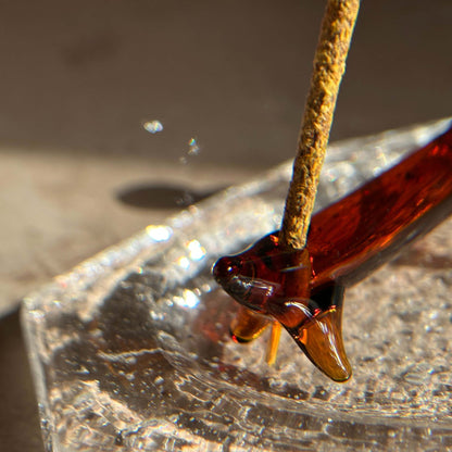 Close-up of a handcrafted apple dog incense holder with a bamboo incense stick inserted, placed on a clear glass ash catcher.