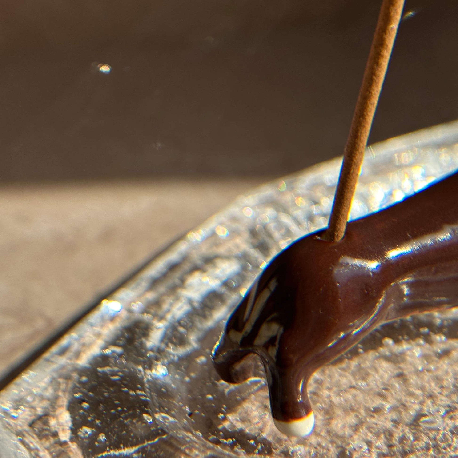 Close-up of a handcrafted ceramic dachshund incense holder holding an incense stick, placed on a textured glass ash catcher.