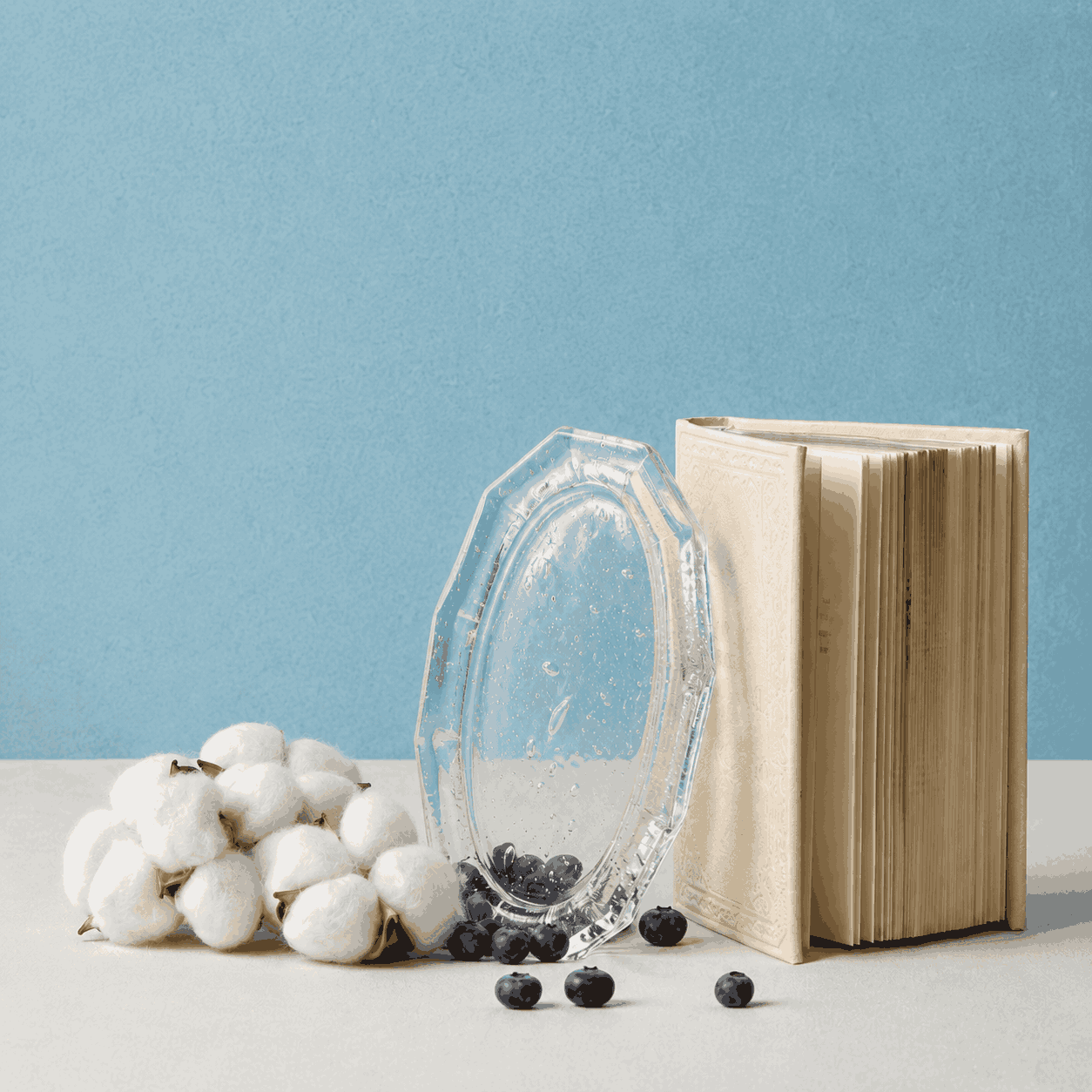 Kumo Glass Incense Tray styled upright beside an open cream-colored book, cotton stems, and scattered blueberries on a white surface with a soft blue background, highlighting the tray’s clear handcrafted glass texture and decorative form.