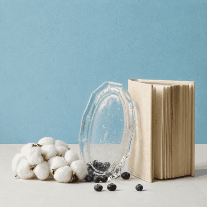 Kumo Glass Incense Tray styled upright beside an open cream-colored book, cotton stems, and scattered blueberries on a white surface with a soft blue background, highlighting the tray’s clear handcrafted glass texture and decorative form.
