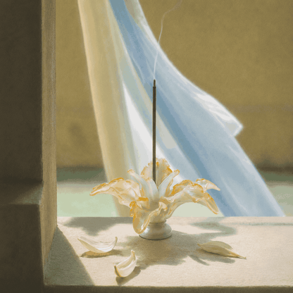 Lily Glass Incense Holder shown on a sunlit windowsill with a burning incense stick, featuring a handcrafted glass incense holder shaped like a blooming lily with softly curved petals, scattered white petals, and gentle smoke.
