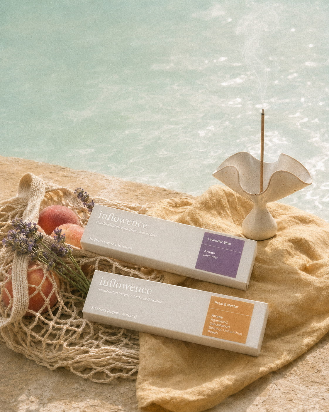 Inflowence Lavender Bliss lavender incense box and Petal & Nectar flower incense box arranged by the pool on soft yellow fabric, with a white ceramic incense holder, burning incense stick, lavender sprigs, peaches in woven netting, textured stone, calm blue water, and gentle summer light.