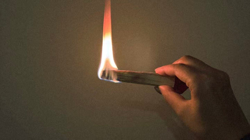A hand holds a burning stick of wood, likely Palo Santo, against a dark background. A bright, orange and white flame is visible at the end of the wood stick, suggesting it has just been lit for the purpose of smudging or releasing its aromatic smoke for purification, relaxation, or spiritual cleansing.