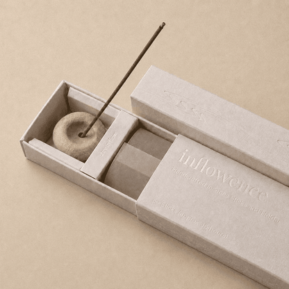 Close-up of an open inflowence incense box with a ceramic holder and a lit incense stick on an orange background.