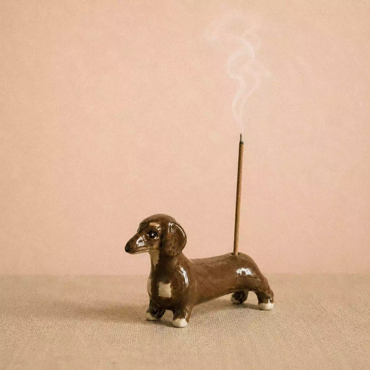 Sausage Dog Incense Holder, a playful hand-painted ceramic incense holder shaped like a dachshund, shown on a soft pink background with a burning incense stick and gentle smoke.