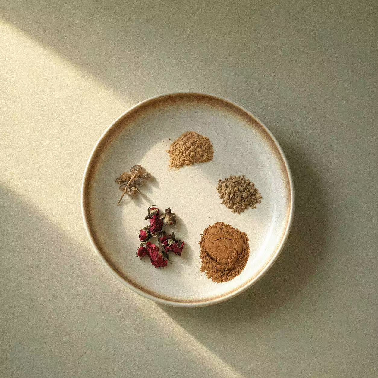 Natural ingredients used in Inflowence incense, shown on a ceramic plate with dried rose buds, resin, wood powders, and botanical materials in soft natural light, highlighting the plant-based elements behind the incense blends.