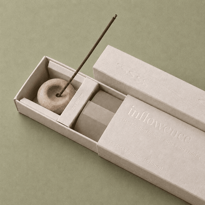 Close-up of an open inflowence incense box with a ceramic holder and a lit incense stick on a green background.
