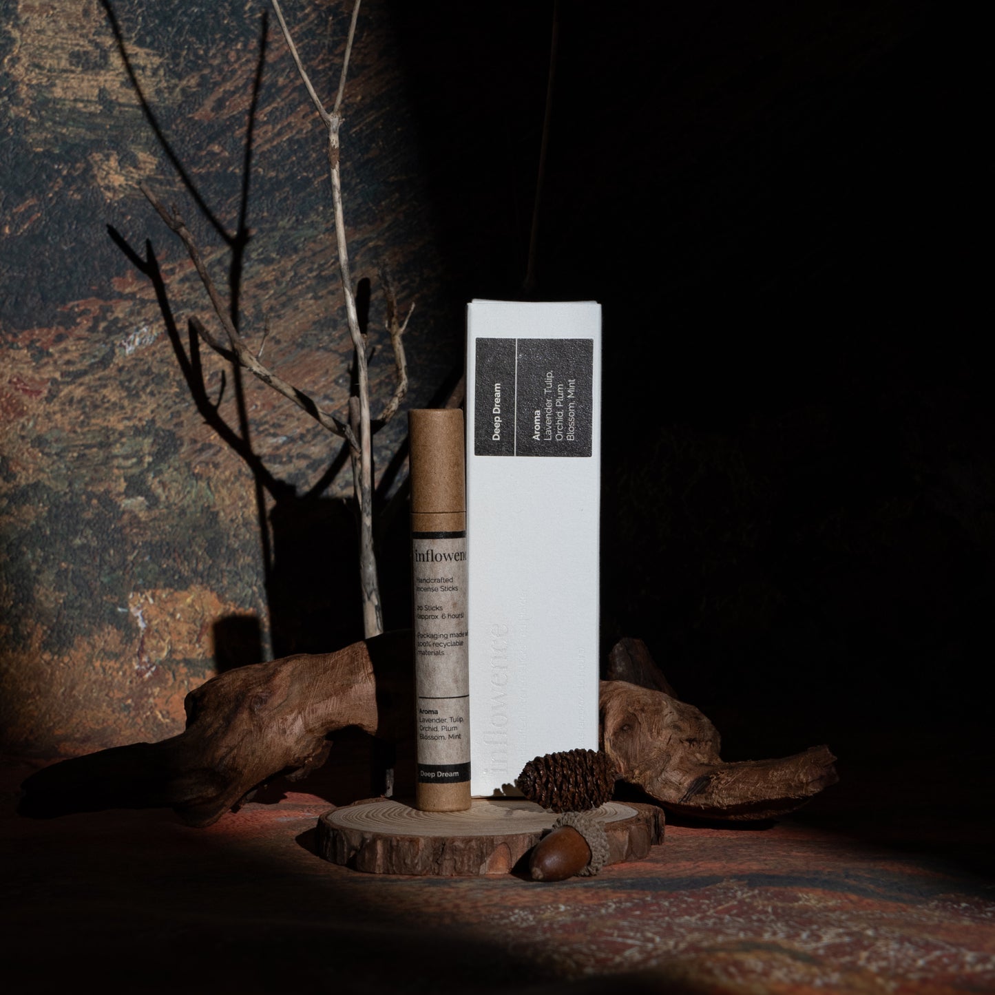Deep Dream incense box and tube set displayed with wood and branches, in front of a green-yellow-red oil painting canvas. Dappled light and shadow highlight the products, evoking a warm, tranquil evening ritual.