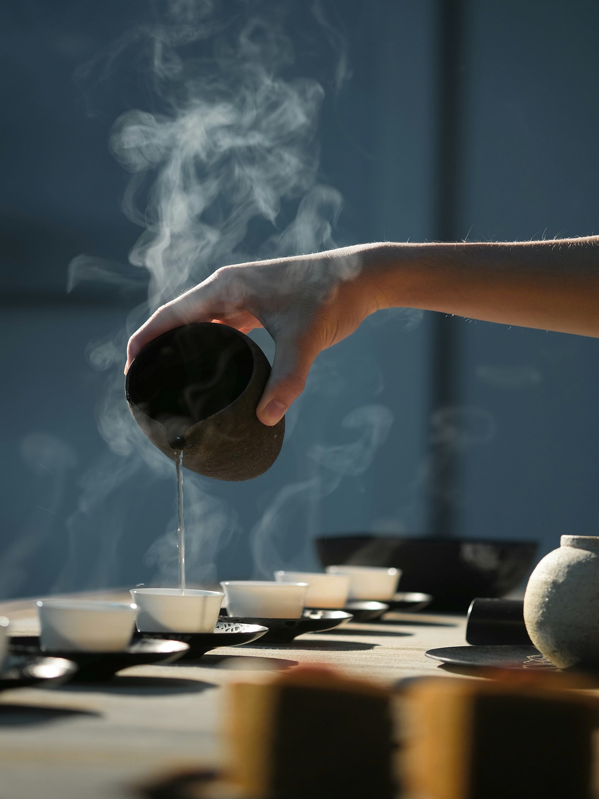 A hand pouring tea from a large clay pot into one of several small cups arranged on the ground, with visible steam rising—capturing warmth, ritual, and grounding presence.