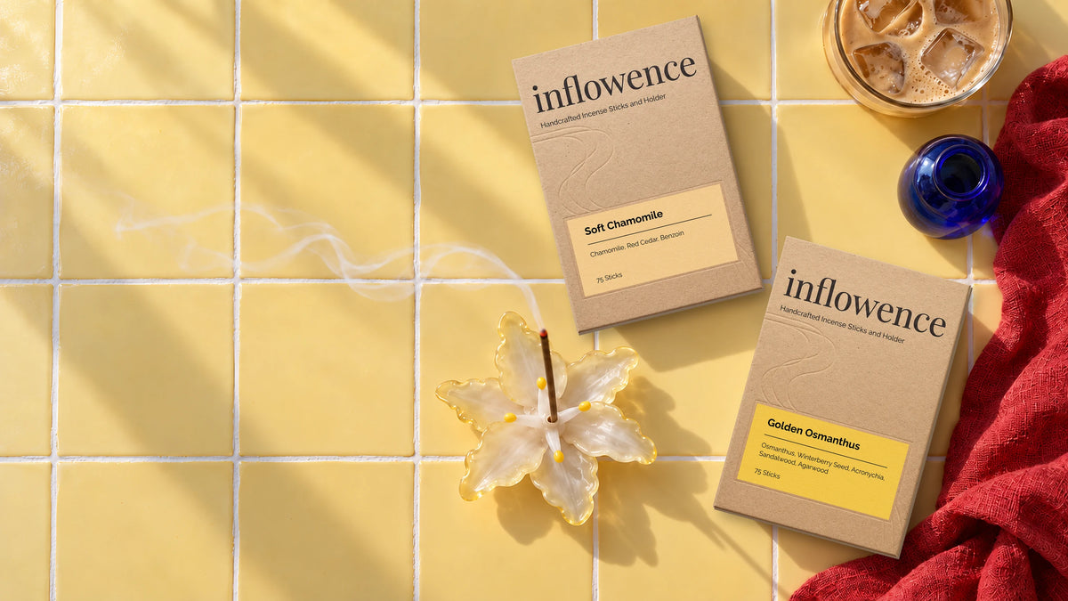 inflowence Soft Chamomile and Golden Osmanthus incense boxes on sunlit yellow tiles, with a lit incense stick in a glass flower holder releasing soft smoke beside iced coffee