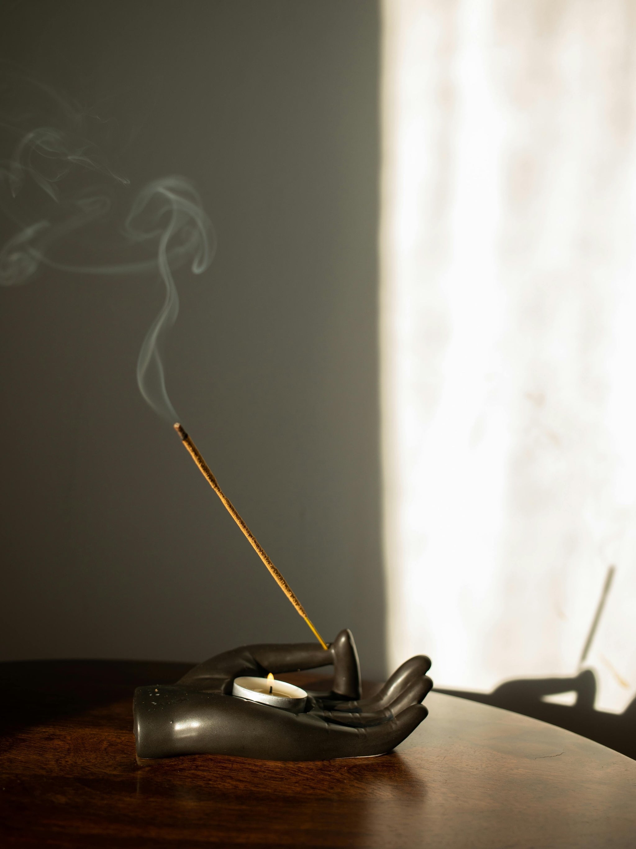 Burning incense stick in a hand-shaped holder on a table, with soft shadows and warm light—symbolizing ritual and calm atmosphere.