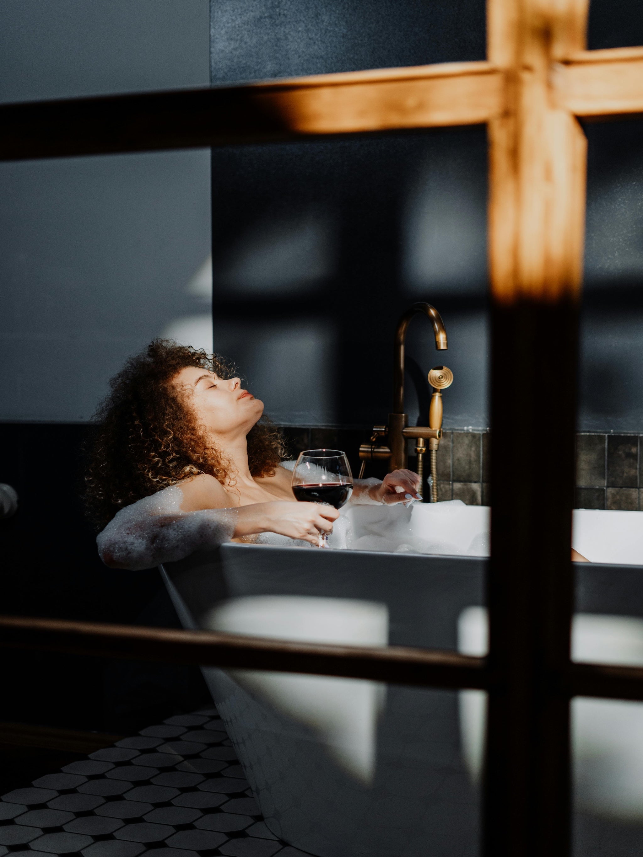 A woman with curly hair holding a glass of wine in a detached bubble bath tub on a patterned tile floor, with soft window shadows cast on the wall—capturing a quiet, indulgent moment of rest.