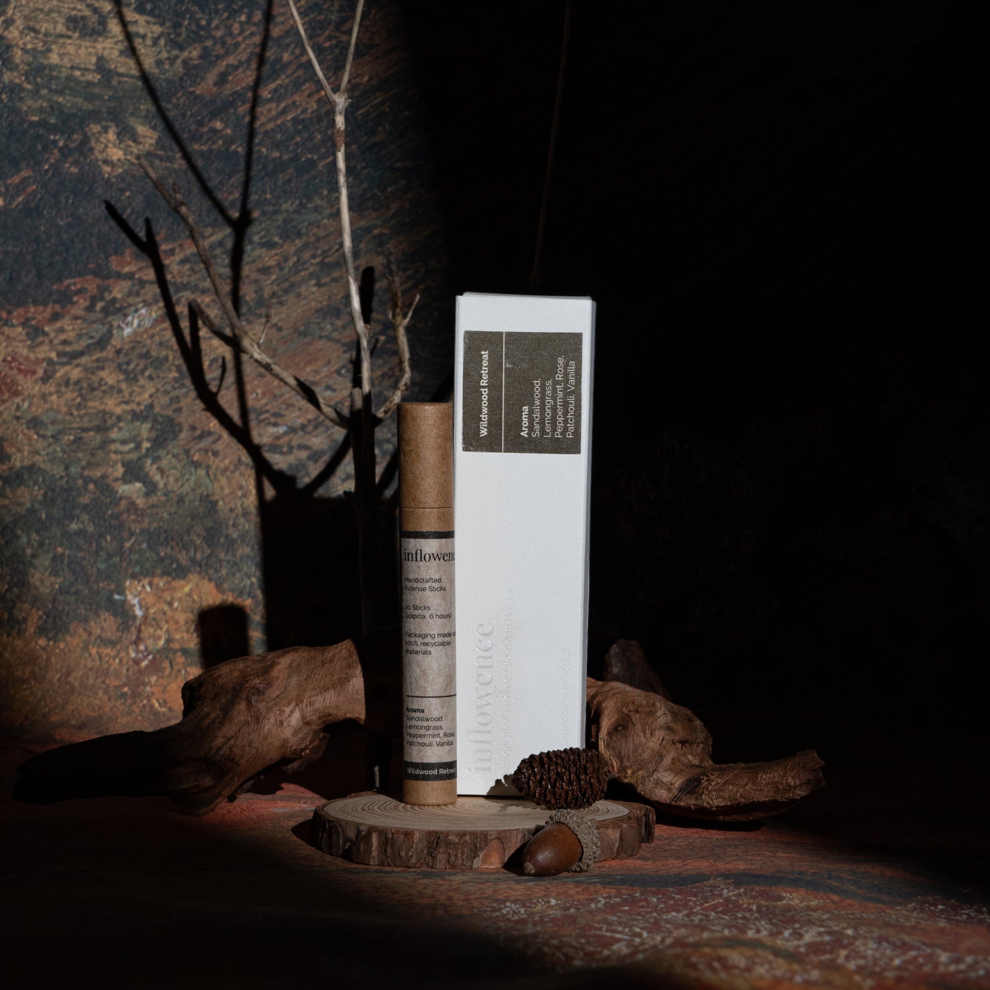 Wildwood Retreat incense box and tube set displayed with wood and branches, in front of a green-yellow-red oil painting canvas. Dappled light and shadow highlight the products, evoking a warm, tranquil evening ritual.