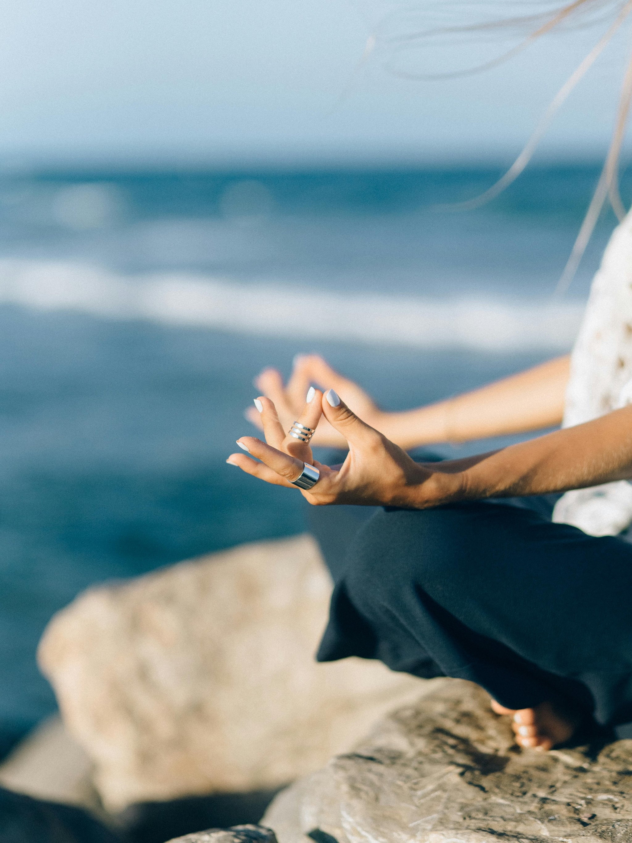 Woman meditating on a rock in front of a blue sea, with the wind gently blowing her hair, embodying calm, presence, and connection with nature.